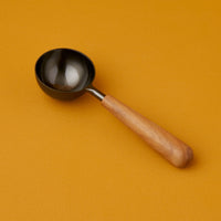 A black stainless steel scoop with a teak wood handle on an orange background.
